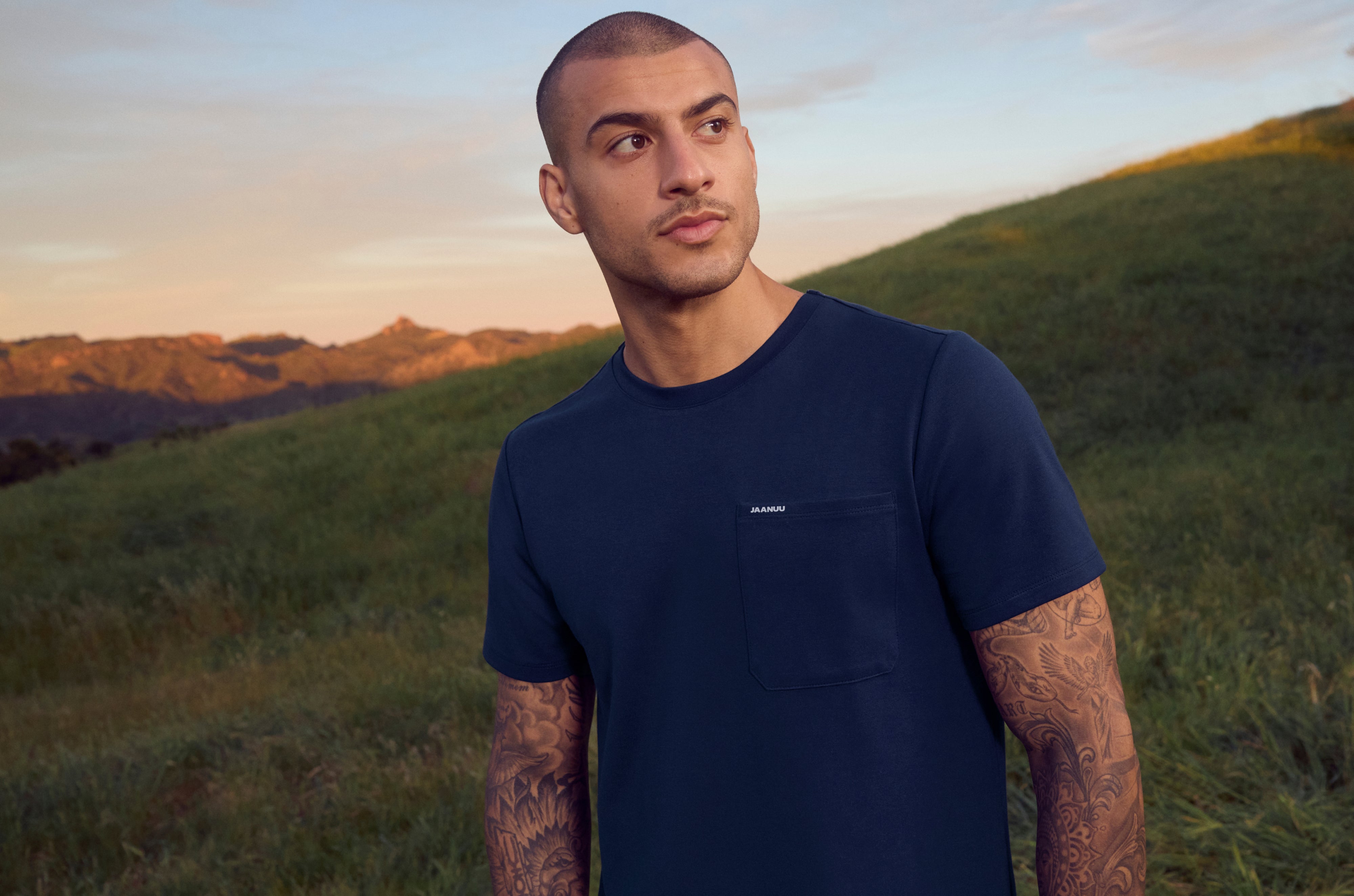 Man in a navy blue Jaanuu crewneck pocket tee standing on a grassy hillside at sunset, with mountains behind him