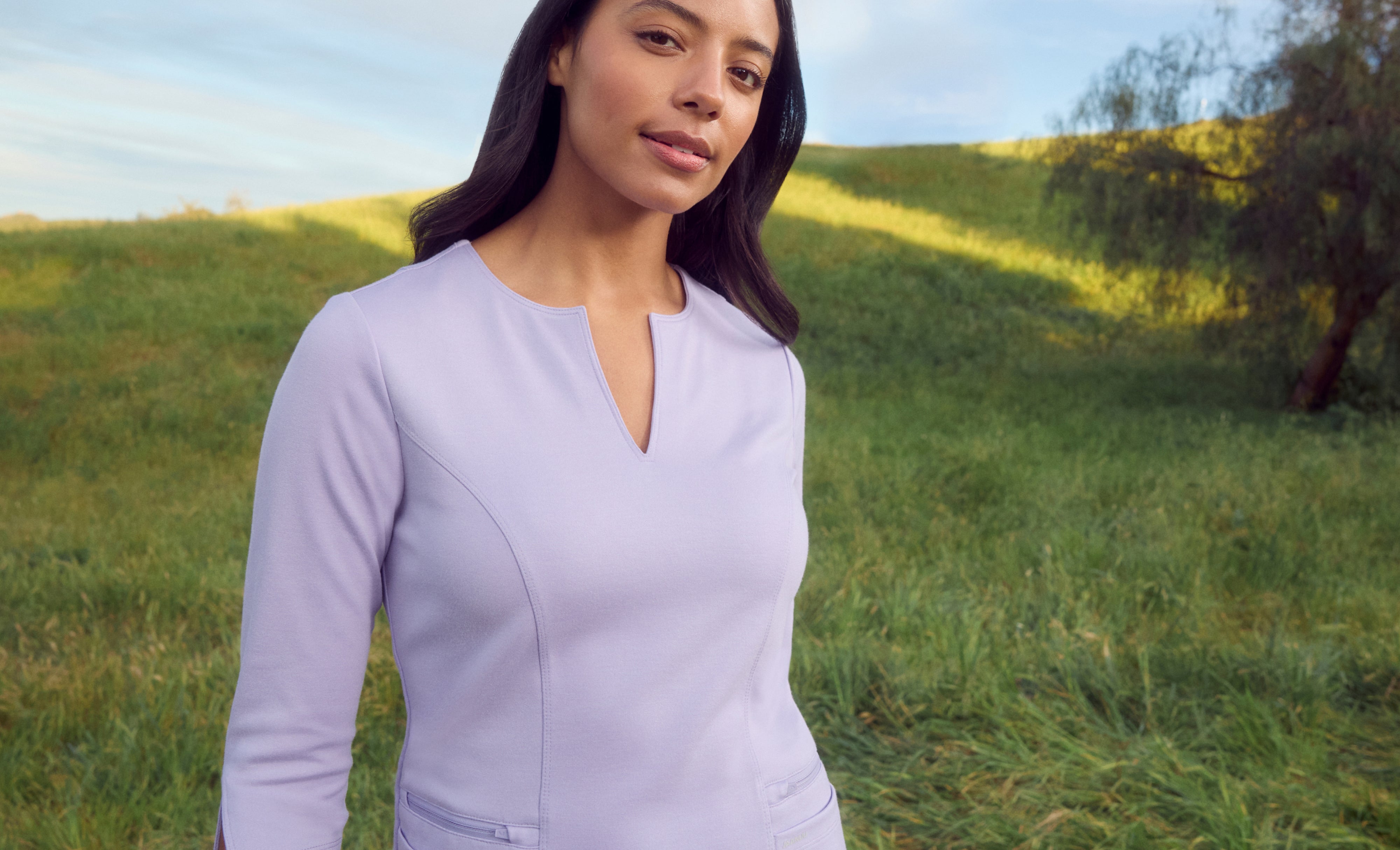 Woman in a light purple Jaanuu notch-neck scrub top with three-quarter sleeves and zip pockets, standing on a grassy hillside with a tree behind her
