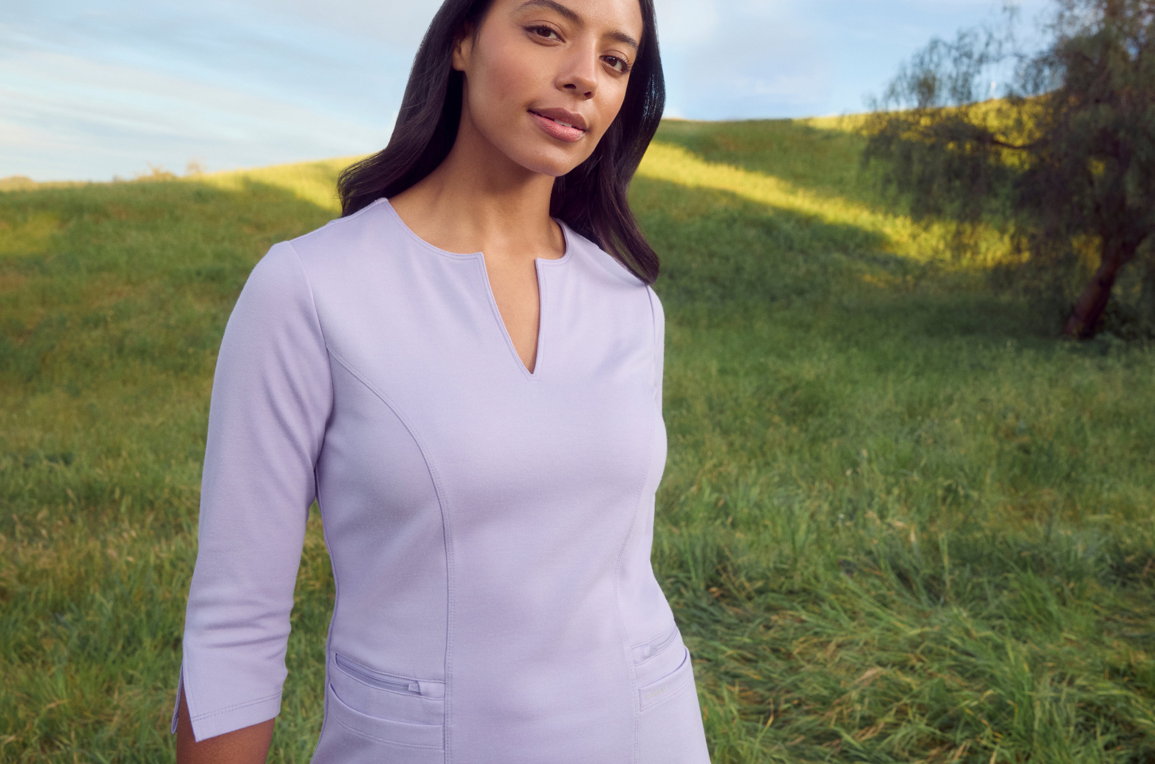 Woman in a light purple Jaanuu notch-neck scrub top with three-quarter sleeves and zip pockets, standing on a grassy hillside with a tree behind her