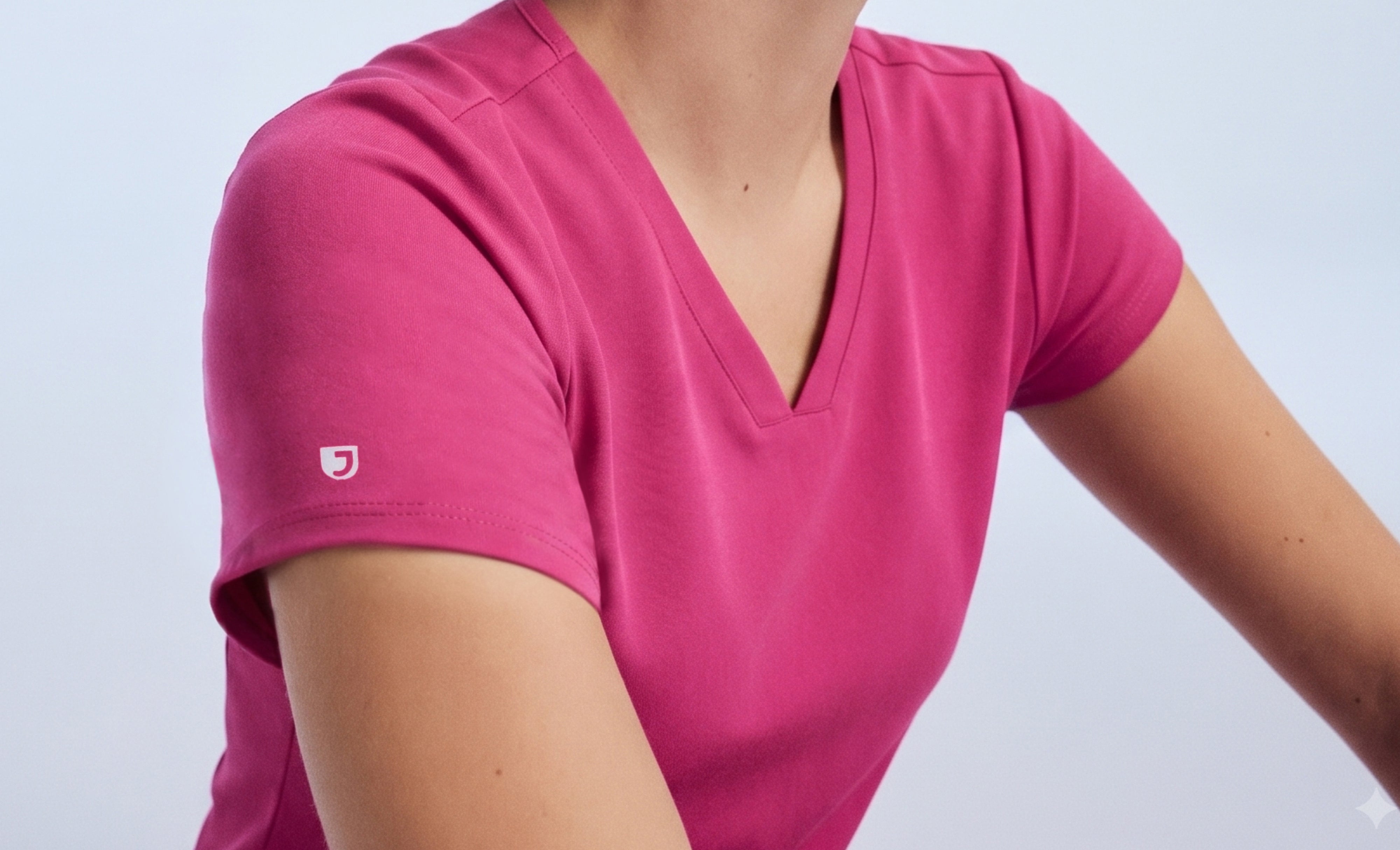 Close-up of a woman's shoulder and V-neckline in a Warrior Pink (bright pink) Jaanuu scrub top with the white Jaanuu J logo on the sleeve, against a pale blue backdrop