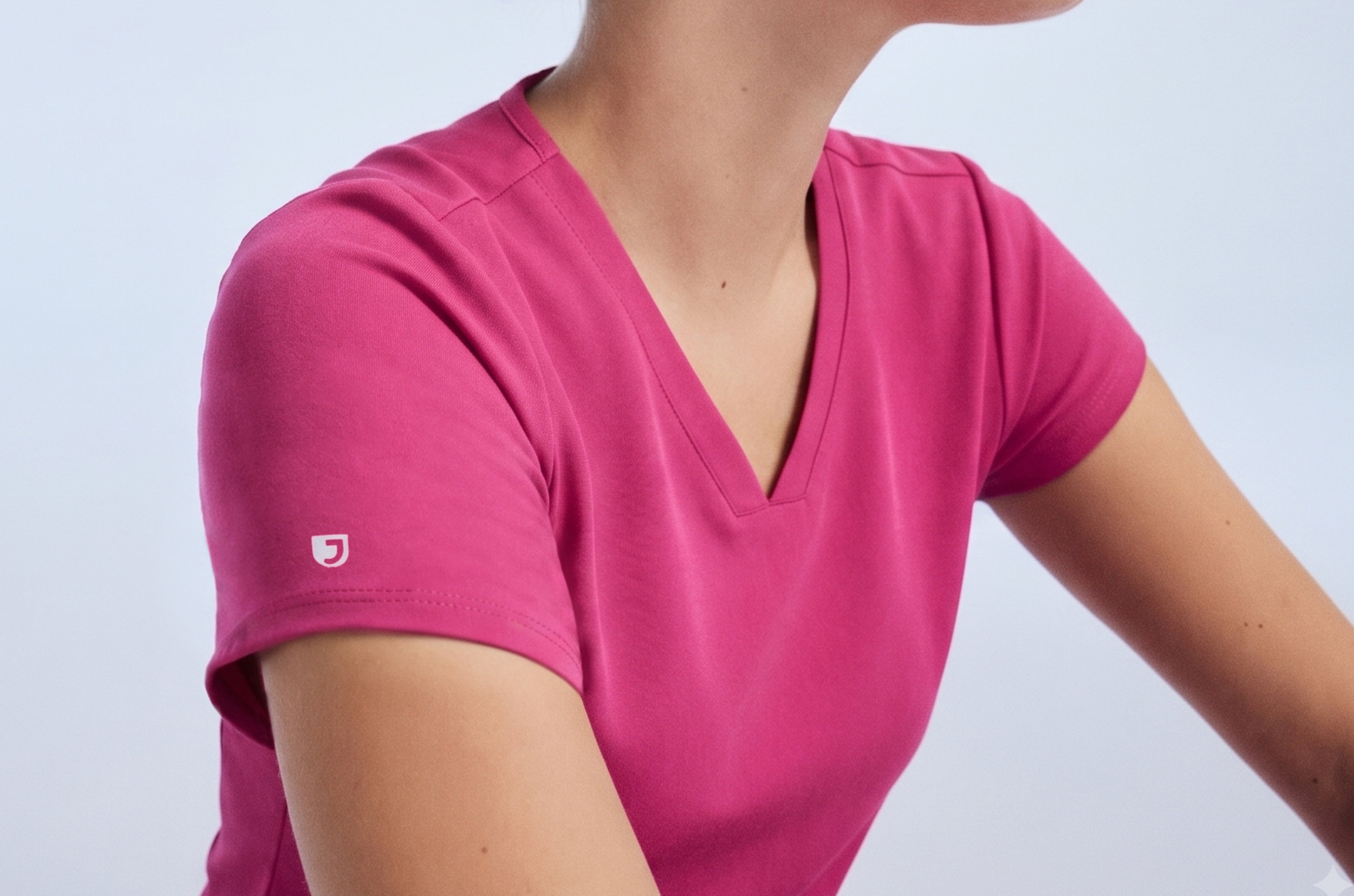 Close-up of a woman's shoulder and V-neckline in a Warrior Pink (bright pink) Jaanuu scrub top with the white Jaanuu J logo on the sleeve, against a pale blue backdrop