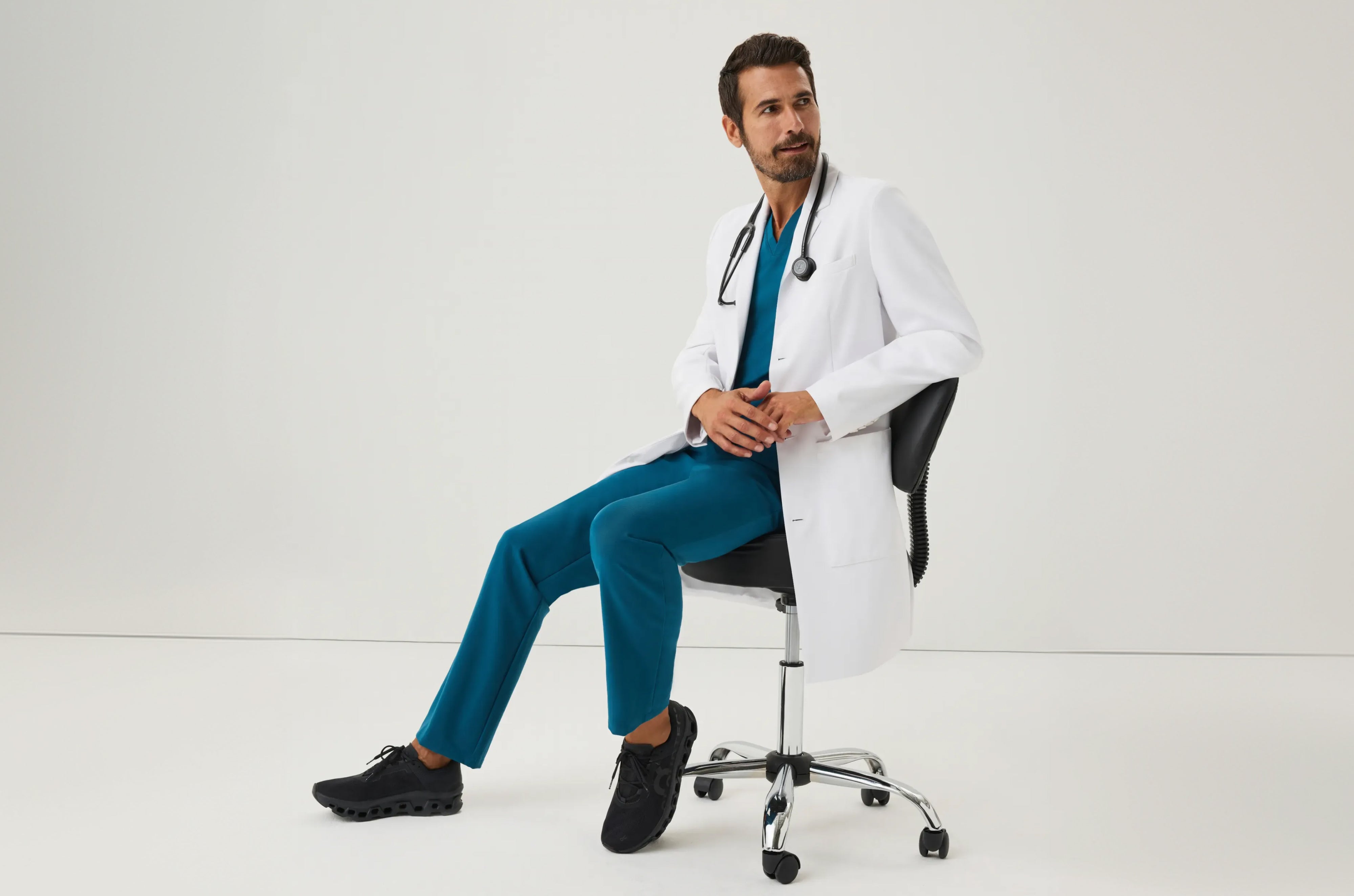 Man in a Jaanuu white coat over a teal V-neck scrub top and scrub pants with a stethoscope around his neck, sitting on a rolling stool against a white backdrop