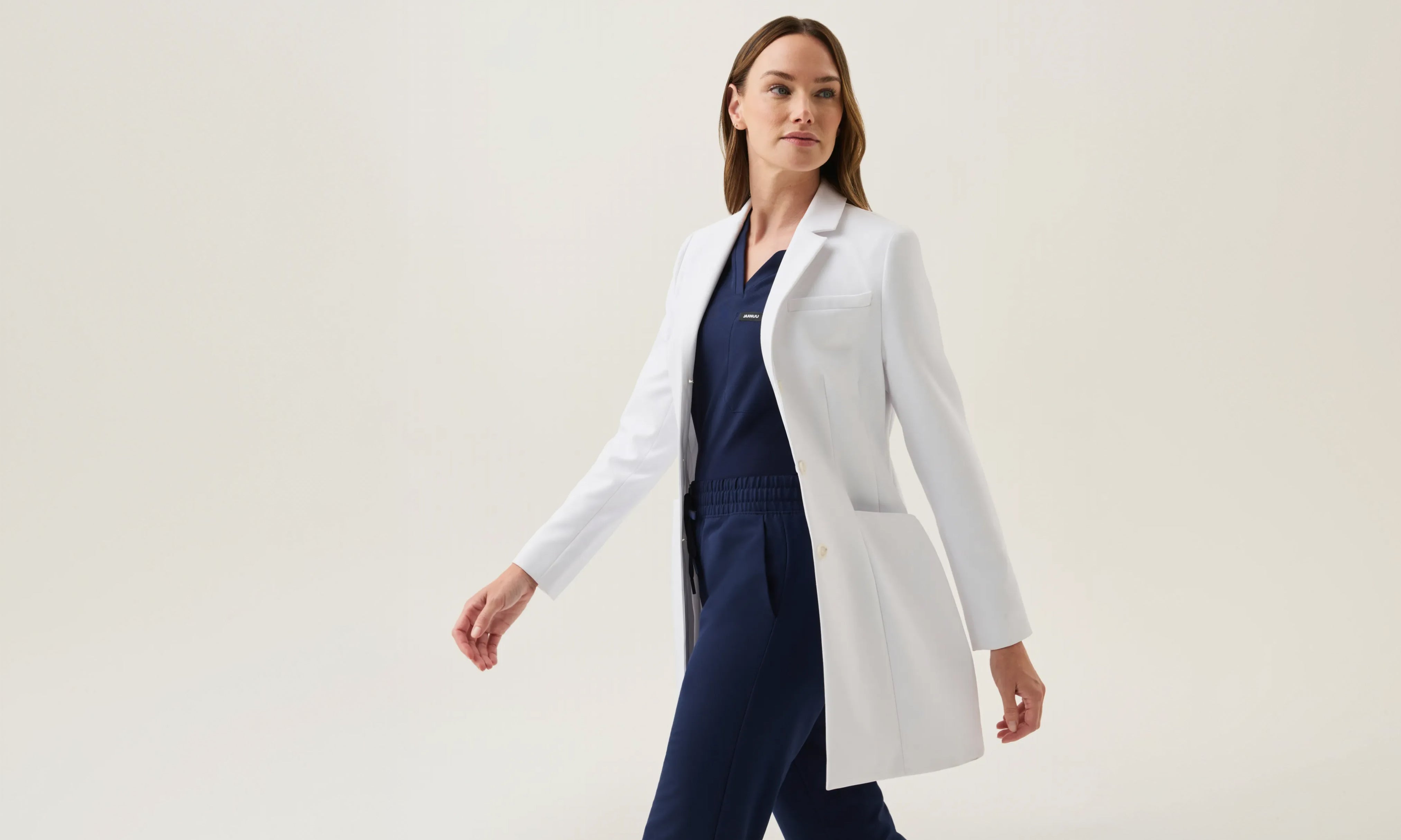 Woman in a Jaanuu white coat over a navy blue V-neck scrub top and drawstring scrub pants, walking against a light backdrop