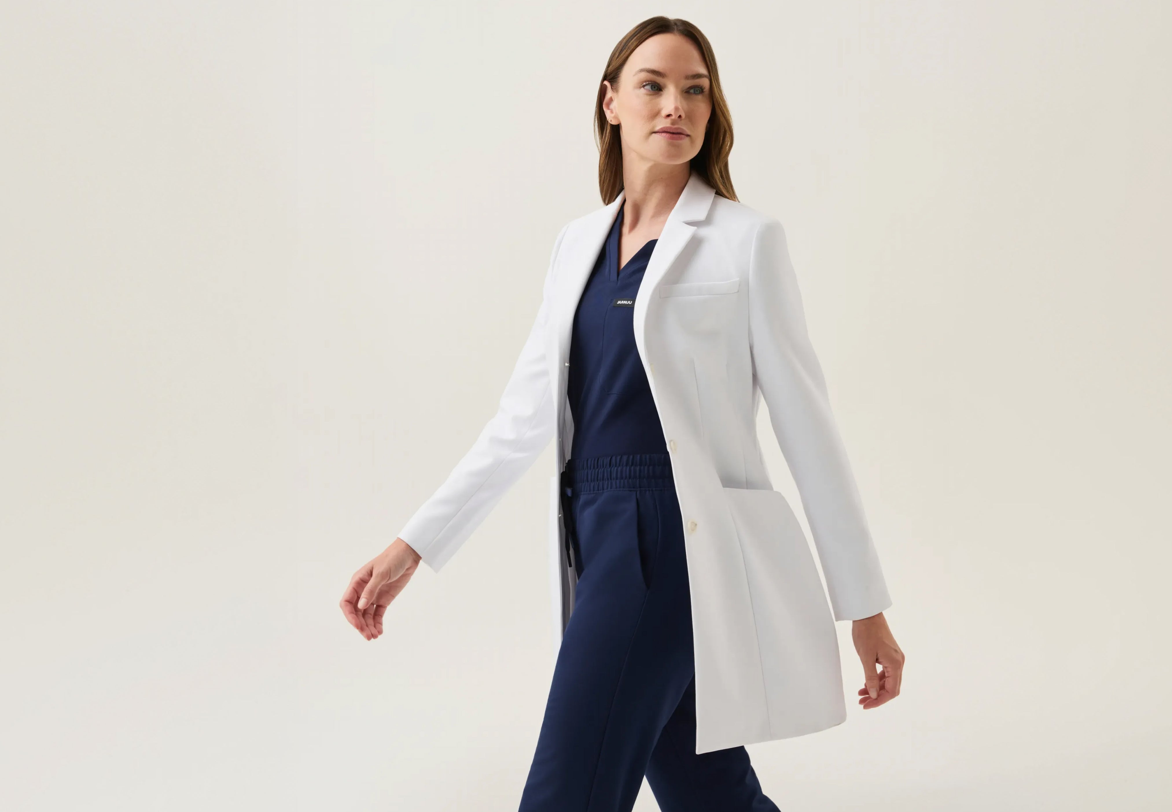 Woman in a Jaanuu white coat over a navy blue V-neck scrub top and drawstring scrub pants, walking against a light backdrop