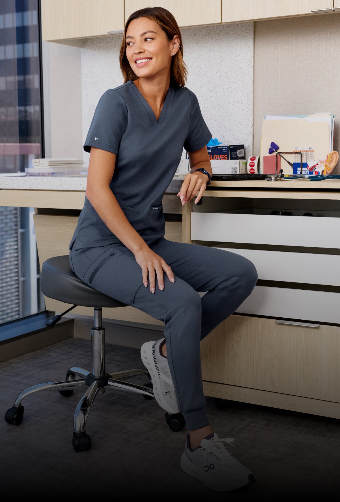 Smiling woman in scrubs seated at a desk in a clinic office