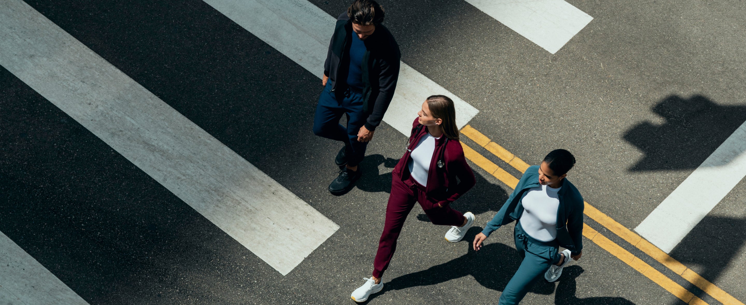 Three students in scrubs and jackets crossing a street, seen from above