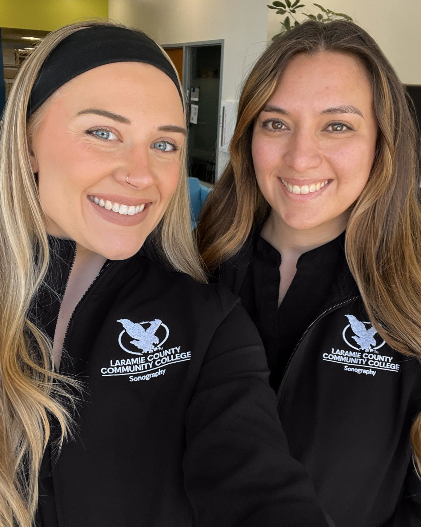 Two smiling students in black jackets with an embroidered college logo taking a selfie