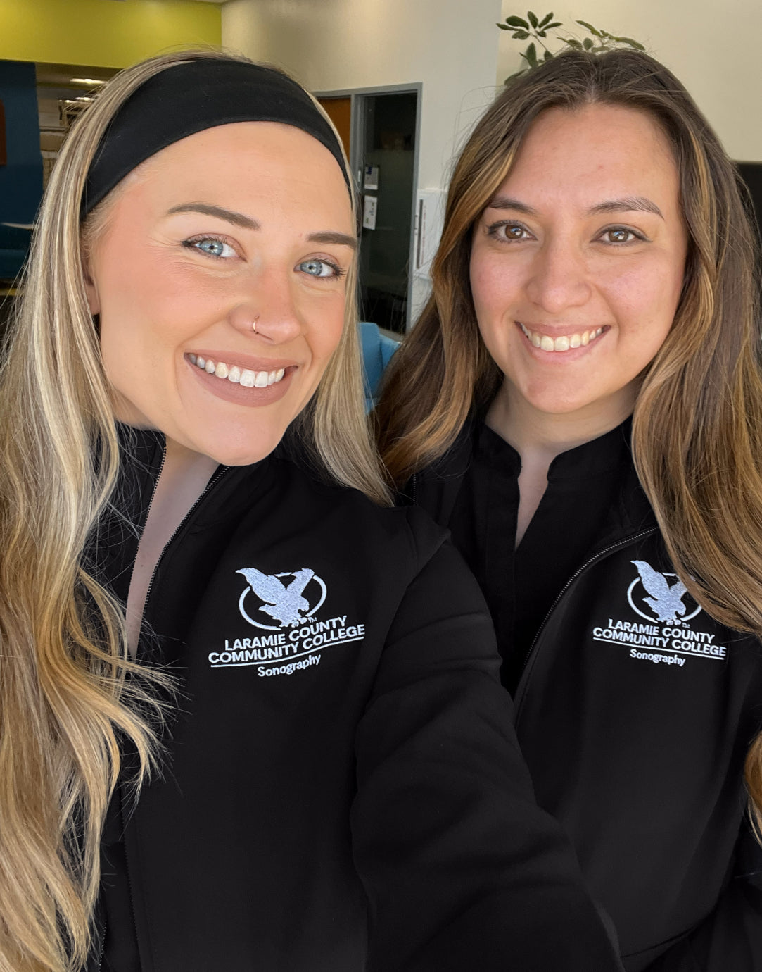 Two smiling students in black jackets with an embroidered college logo taking a selfie
