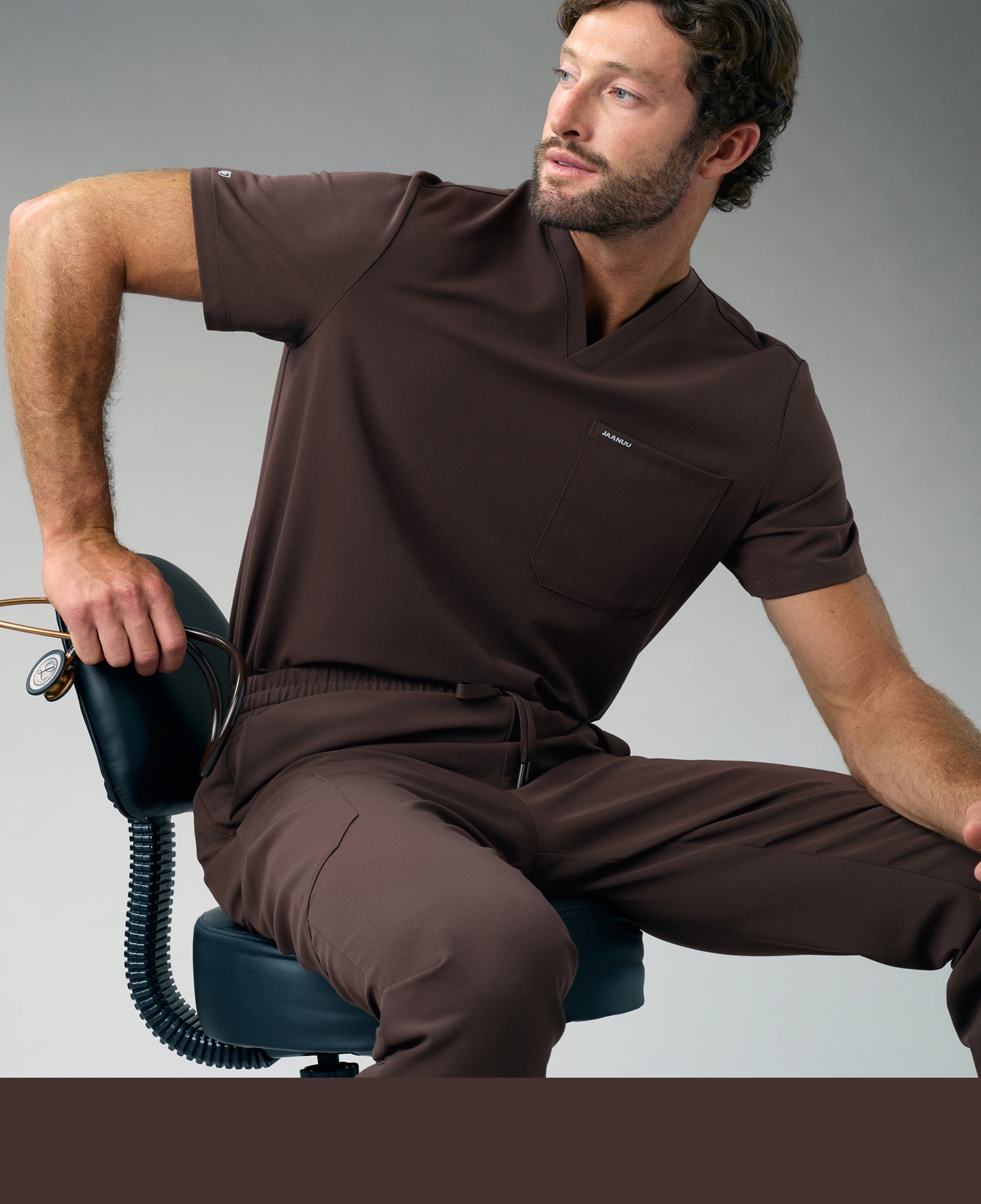 A man in an Espresso Jaanuu V-neck scrub top and scrub pants sits on a black stool holding a stethoscope against its backrest, against a grey background, with an Espresso color band across the bottom