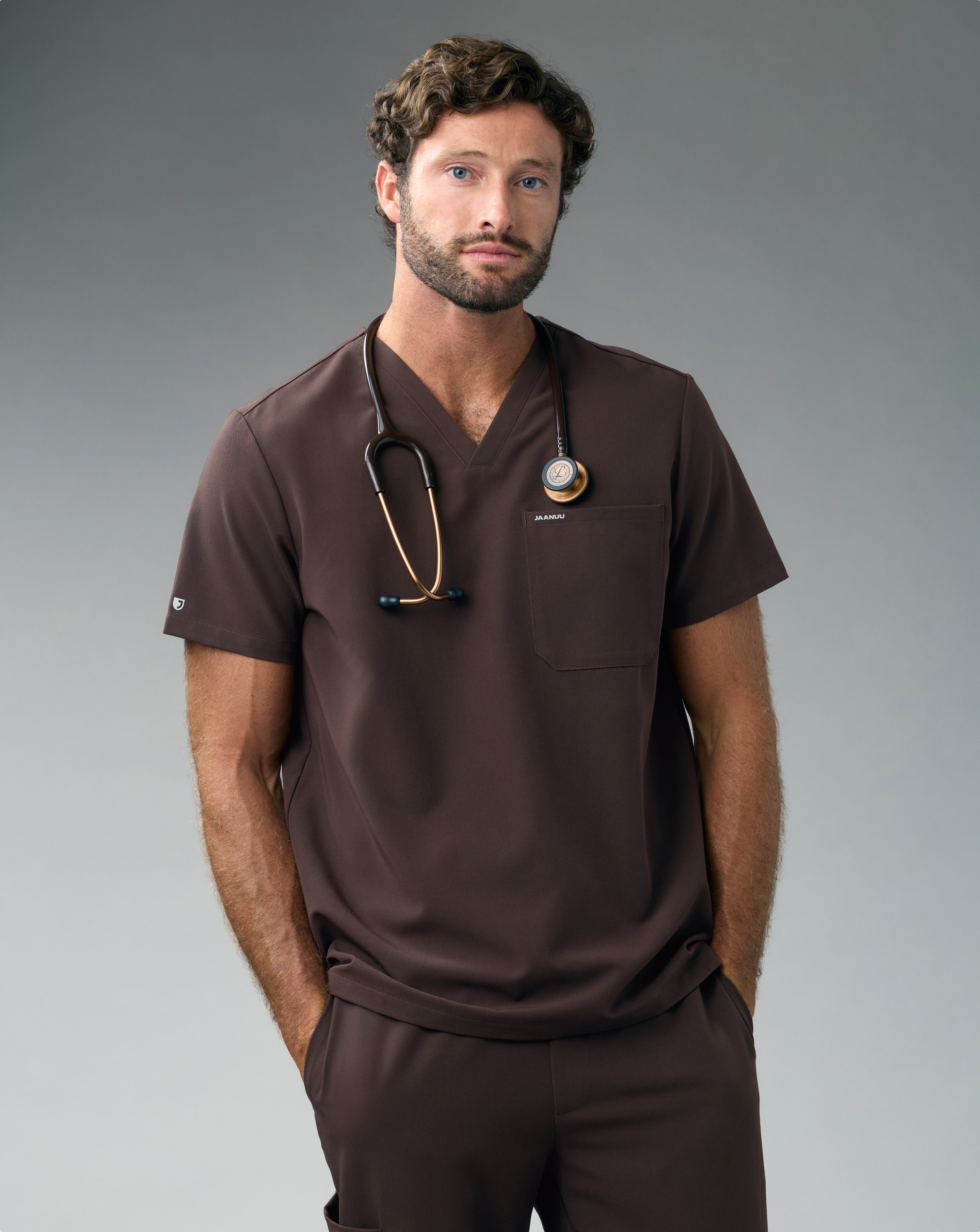 A man in an Espresso Jaanuu scrub top and scrub pants stands with his hands in his pockets and a stethoscope around his neck, against a grey backdrop