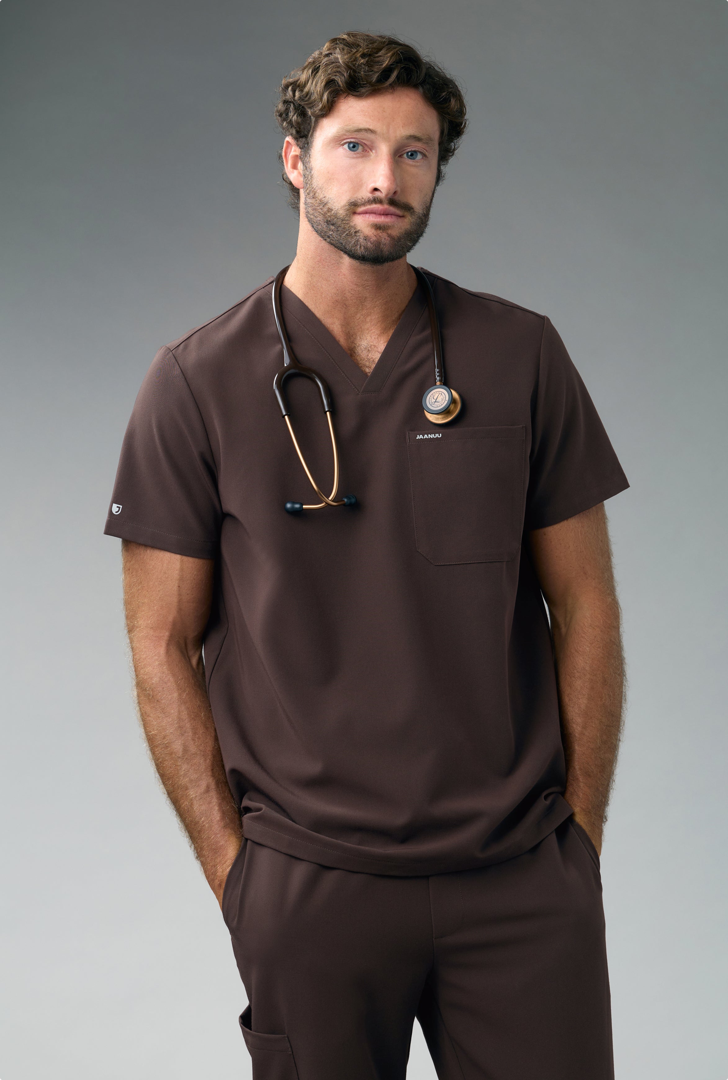A man in an Espresso Jaanuu scrub top and scrub pants stands with his hands in his pockets and a stethoscope around his neck, against a grey backdrop