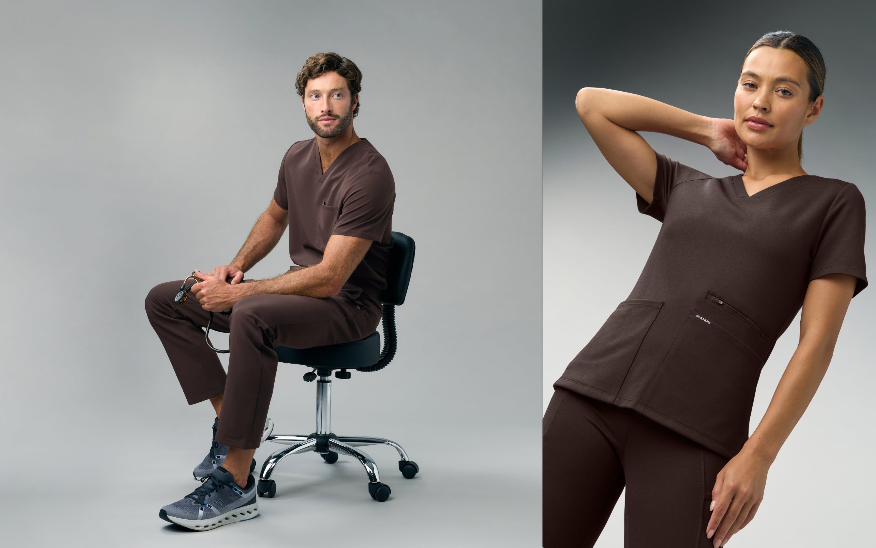 A man in an Espresso Jaanuu scrub top and scrub pants sits on a stool holding a stethoscope, next to a woman in an Espresso Jaanuu V-neck scrub top with one hand behind her neck, against a grey backdrop