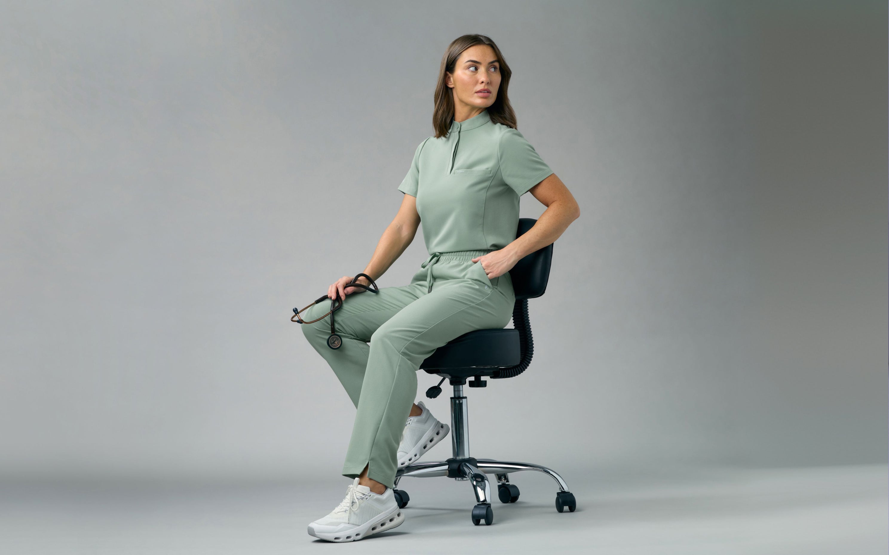 A woman in a Sage Jaanuu stand-collar scrub top and scrub pants sits on a stool holding a stethoscope, against a grey backdrop