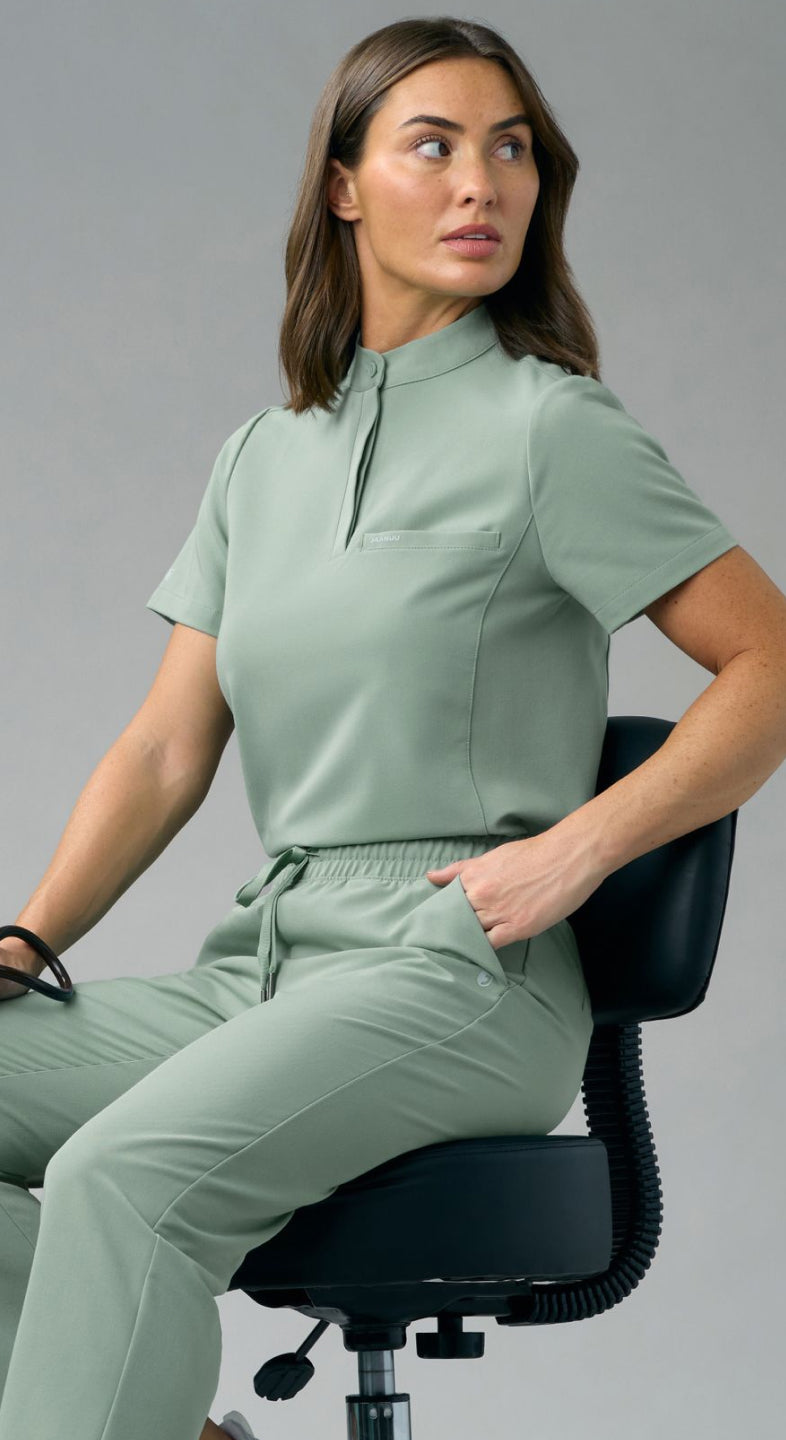 A woman in a Sage Jaanuu stand-collar scrub top and scrub pants sits on a stool holding a stethoscope, against a grey backdrop