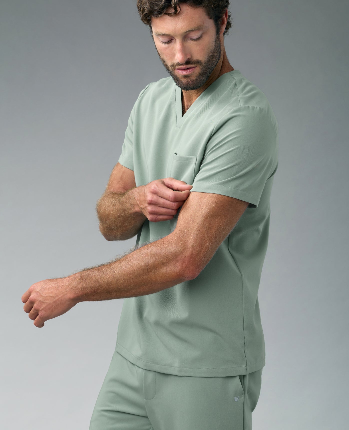 A man in a Sage Jaanuu scrub top and scrub pants looks down and pinches his sleeve, against a grey backdrop