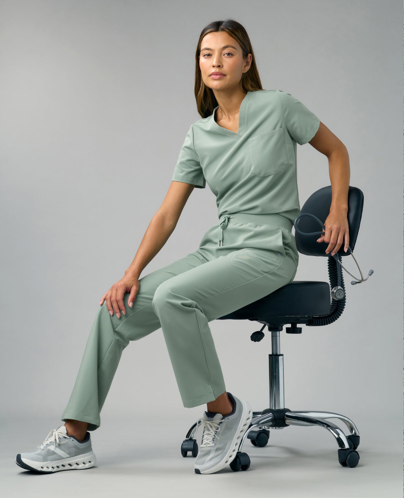 A woman in a Sage Jaanuu scrub top and scrub pants sits on a stool holding a stethoscope, against a grey backdrop