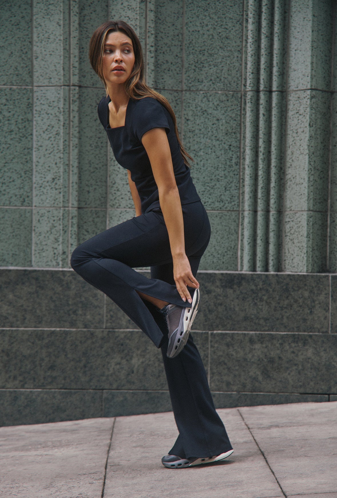 A woman in a Black Jaanuu square-neck scrub top and split-hem flare scrub pants balances on one leg and holds her sneaker, in front of a green stone wall