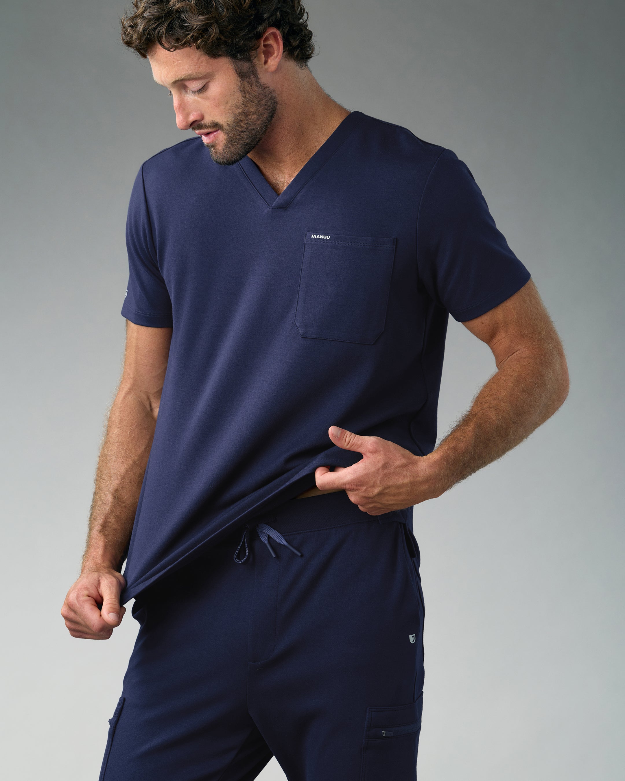 A man in a navy blue Jaanuu V-neck scrub top with a chest pocket and drawstring cargo scrub pants looks down and tugs at the hem of his top, against a grey backdrop