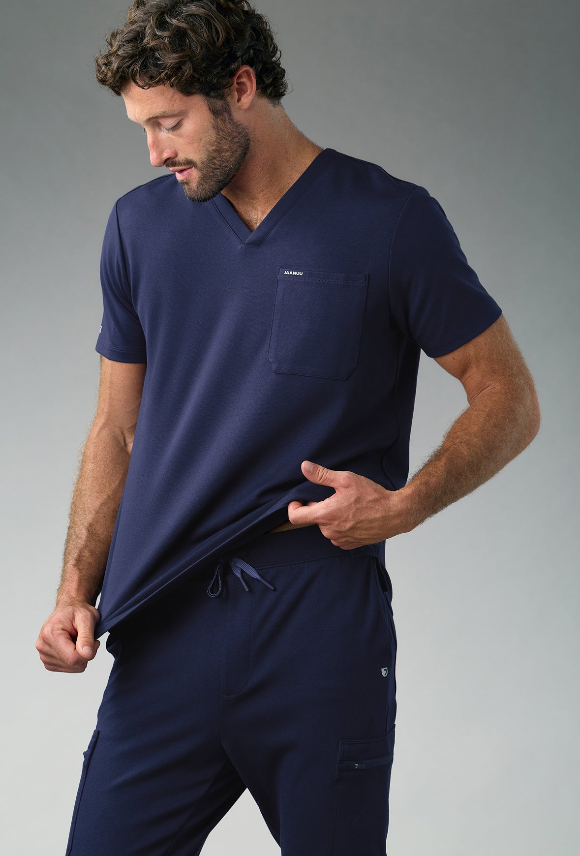A man in a navy blue Jaanuu V-neck scrub top with a chest pocket and drawstring cargo scrub pants looks down and tugs at the hem of his top, against a grey backdrop