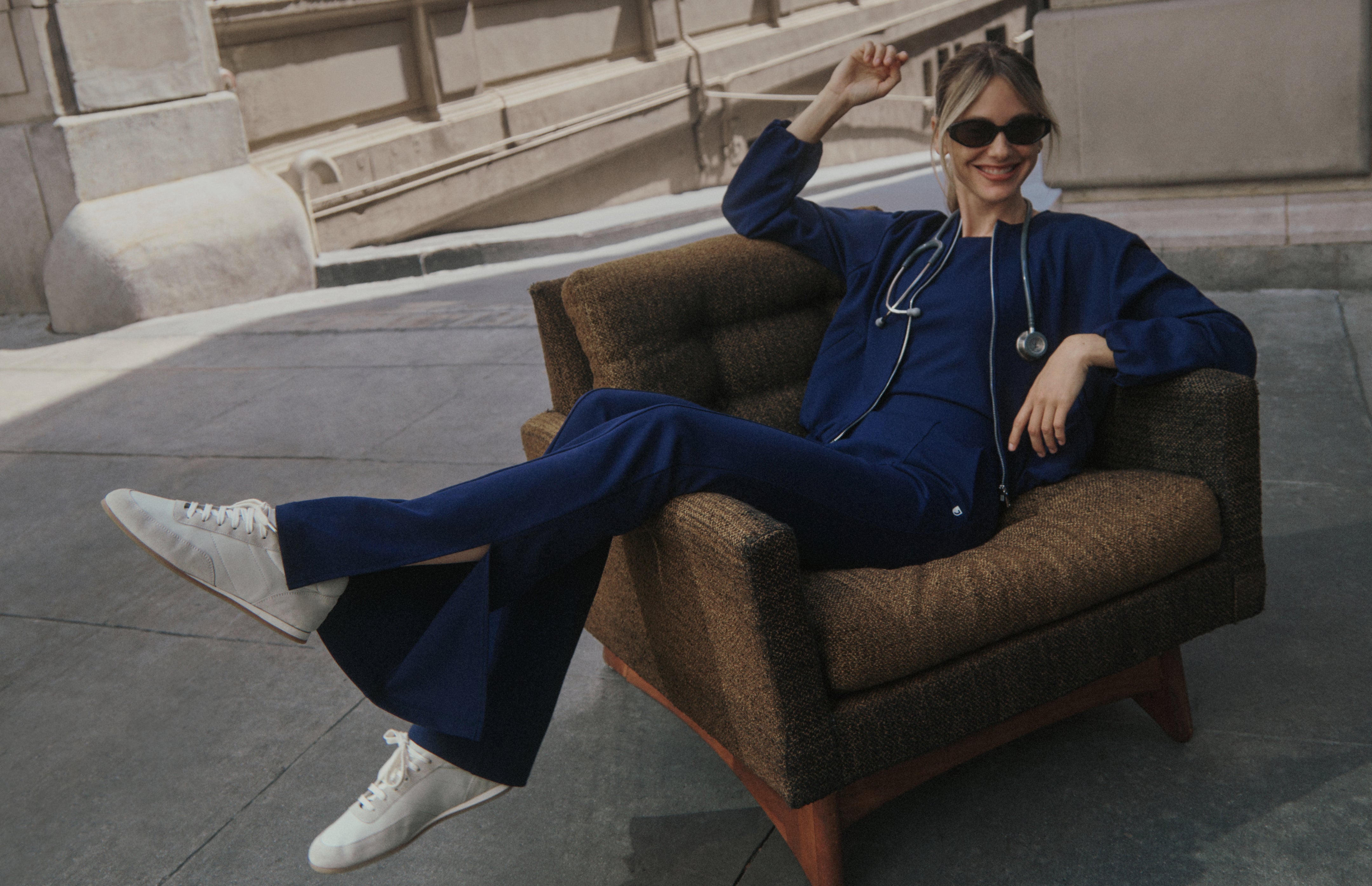 A smiling woman in sunglasses lounges in a brown tweed armchair on a city sidewalk with one arm raised, wearing a Midnight Navy (dark blue) Jaanuu zip-front scrub jacket, scrub top and flare scrub pants with a stethoscope around her neck