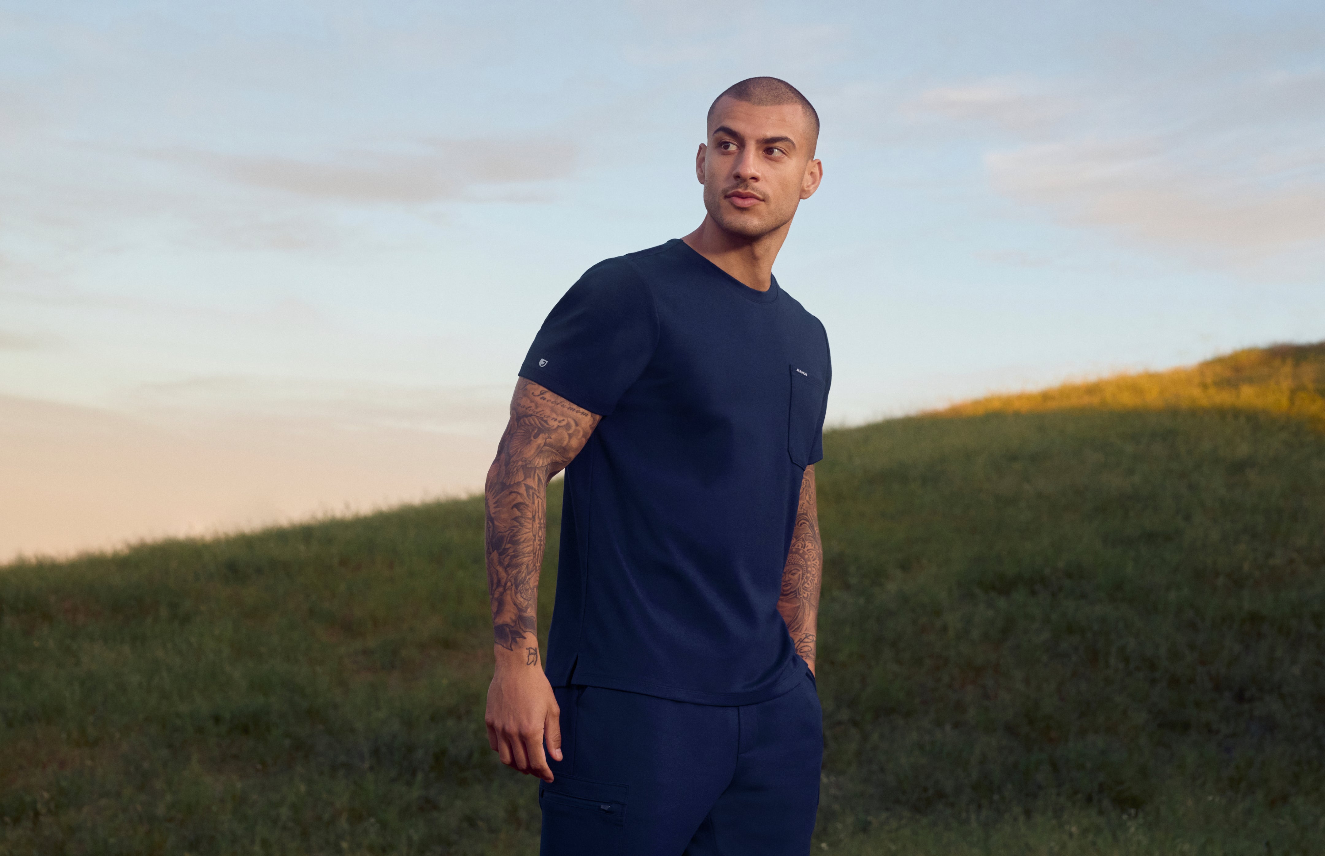 A man in a Midnight Navy (dark blue) Jaanuu crewneck scrub top with a chest pocket and cargo scrub pants stands on a grassy hillside at dusk