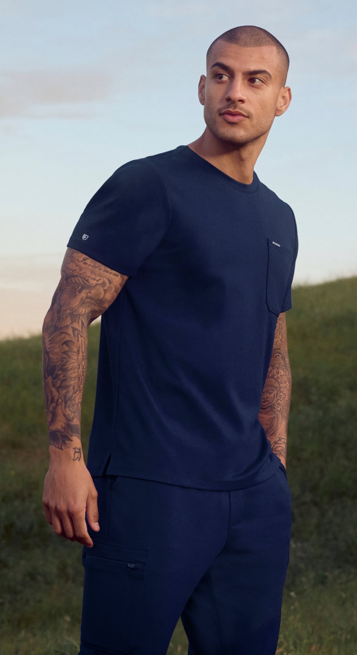 A man in a Midnight Navy (dark blue) Jaanuu crewneck scrub top with a chest pocket and cargo scrub pants stands on a grassy hillside at dusk