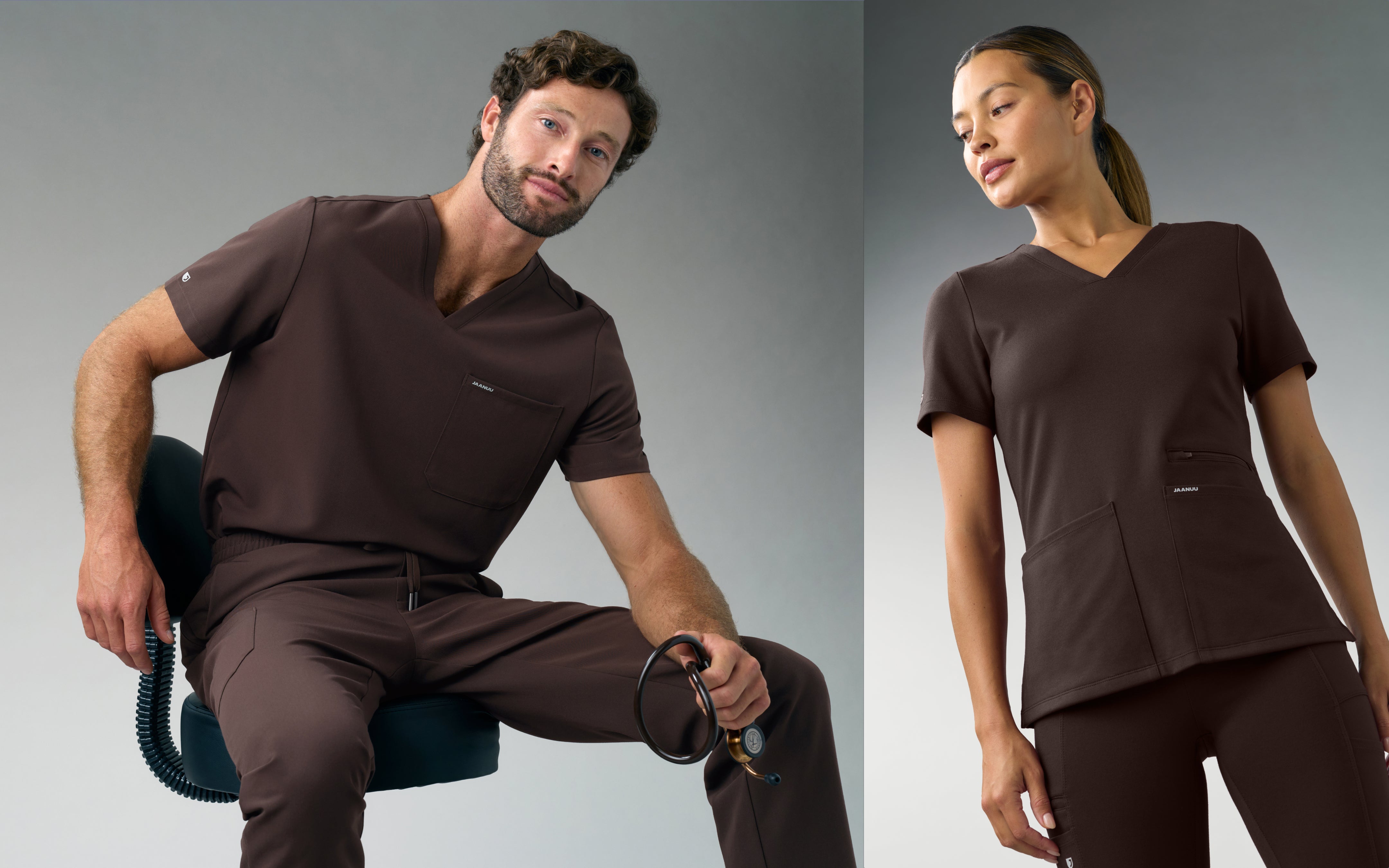 A man in a dark brown Jaanuu V-neck scrub top and scrub pants sits on a stool holding a stethoscope, next to a woman in a dark brown Jaanuu V-neck scrub top and scrub pants, against a grey backdrop