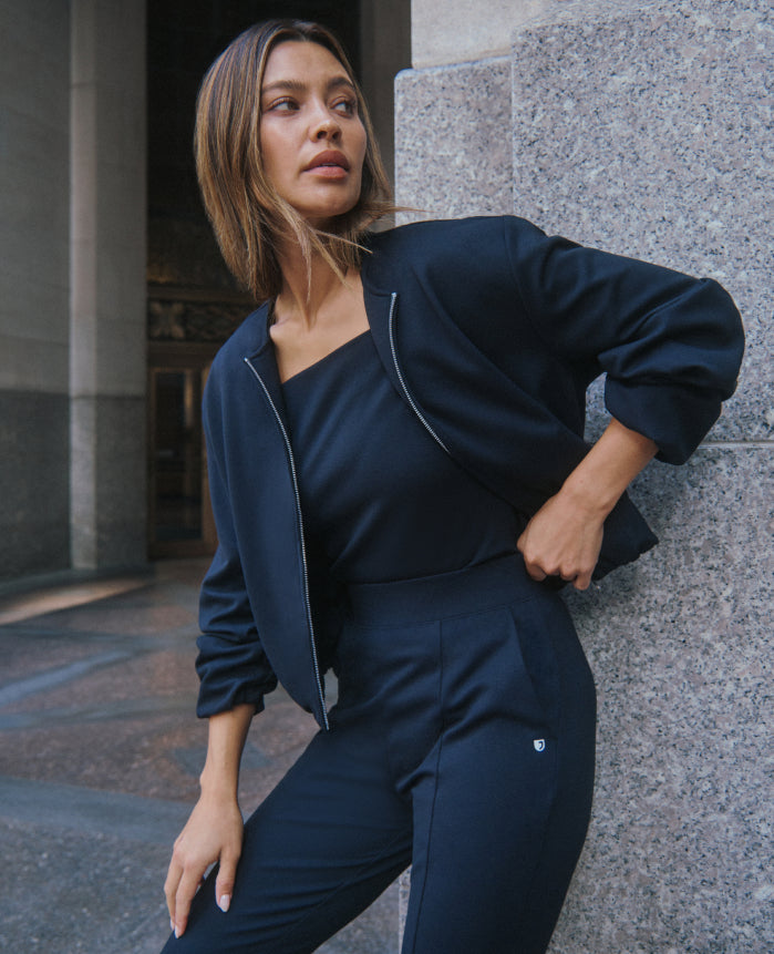 A woman leans against a granite column on a city street in a dark Jaanuu zip-front scrub jacket, square-neck scrub top and scrub pants