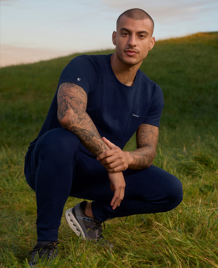 A man crouches on a grassy hillside in a Midnight Navy (dark blue) Jaanuu crewneck scrub top with a chest pocket and jogger scrub pants