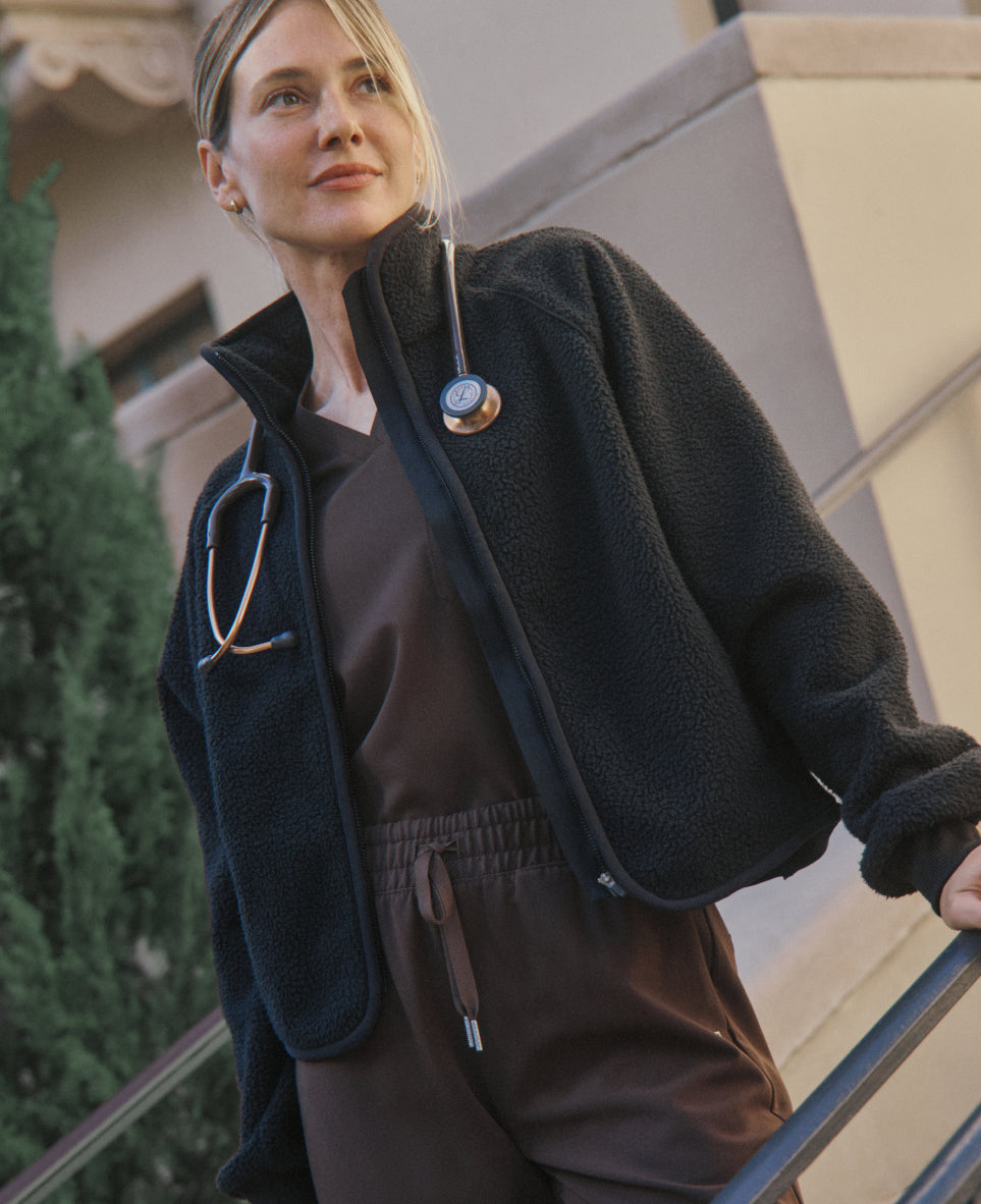 A woman in an open Black Jaanuu sherpa jacket over a dark brown V-neck scrub top and drawstring scrub pants stands on outdoor stairs with a stethoscope around her neck and one hand on the railing