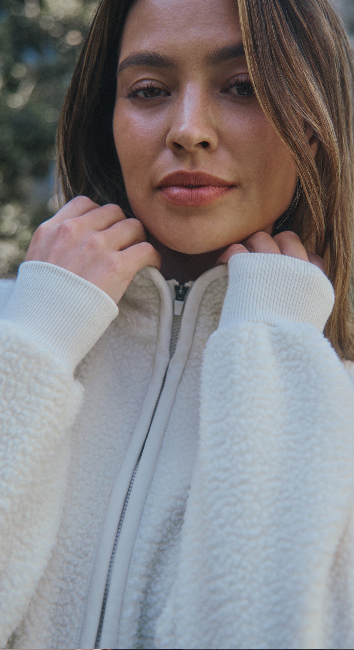 Close-up of a woman holding up the collar of a Birch (cream) Jaanuu zip-front sherpa jacket, with trees blurred behind her