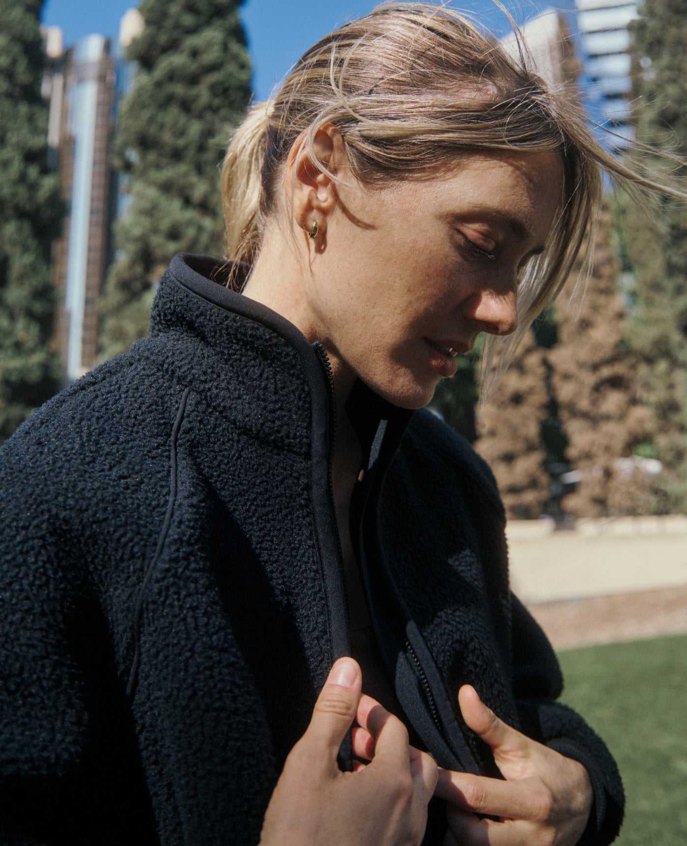 A woman with her hair pulled back looks down as she zips a Black Jaanuu sherpa jacket, with tall trees and city buildings behind her