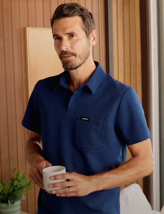 A man wearing a polo scrub top and holding a coffee mug in his hands.